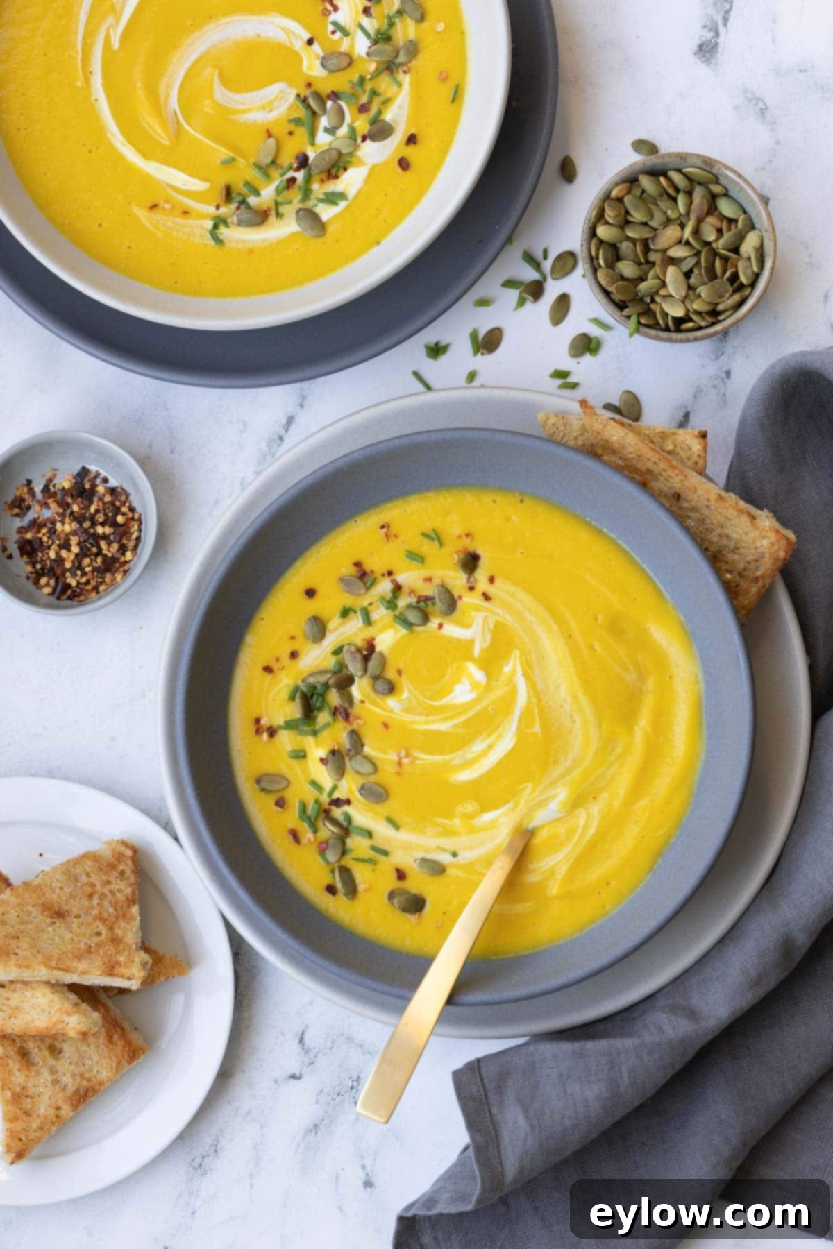 Velvety Butternut Apple Comfort Soup 2 Bright orange butternut squash soup in a gray bowl with garnishes like pumpkin seeds and fresh herbs, served with a gold spoon.