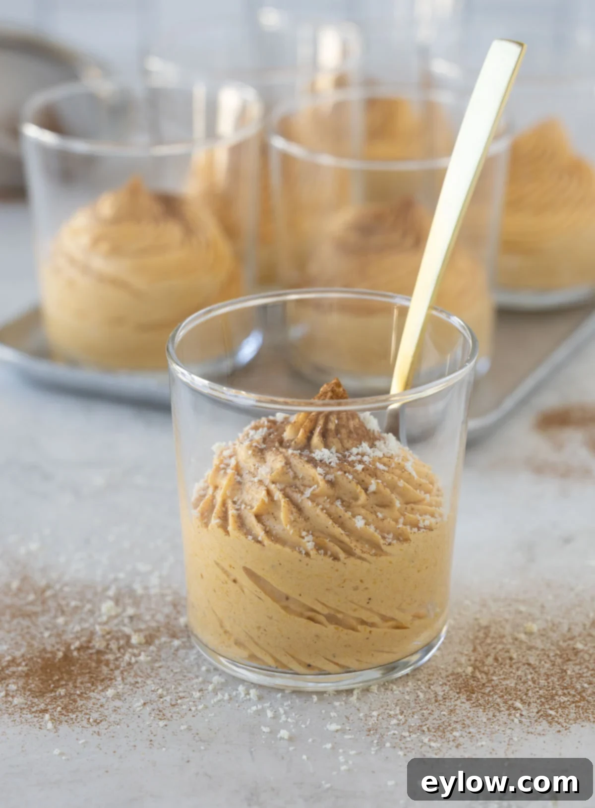 Velvet Pumpkin Mousse No-Bake Treat 2 A glass of creamy pumpkin mousse with a spoon and cinnamon, garnished with white chocolate shavings.