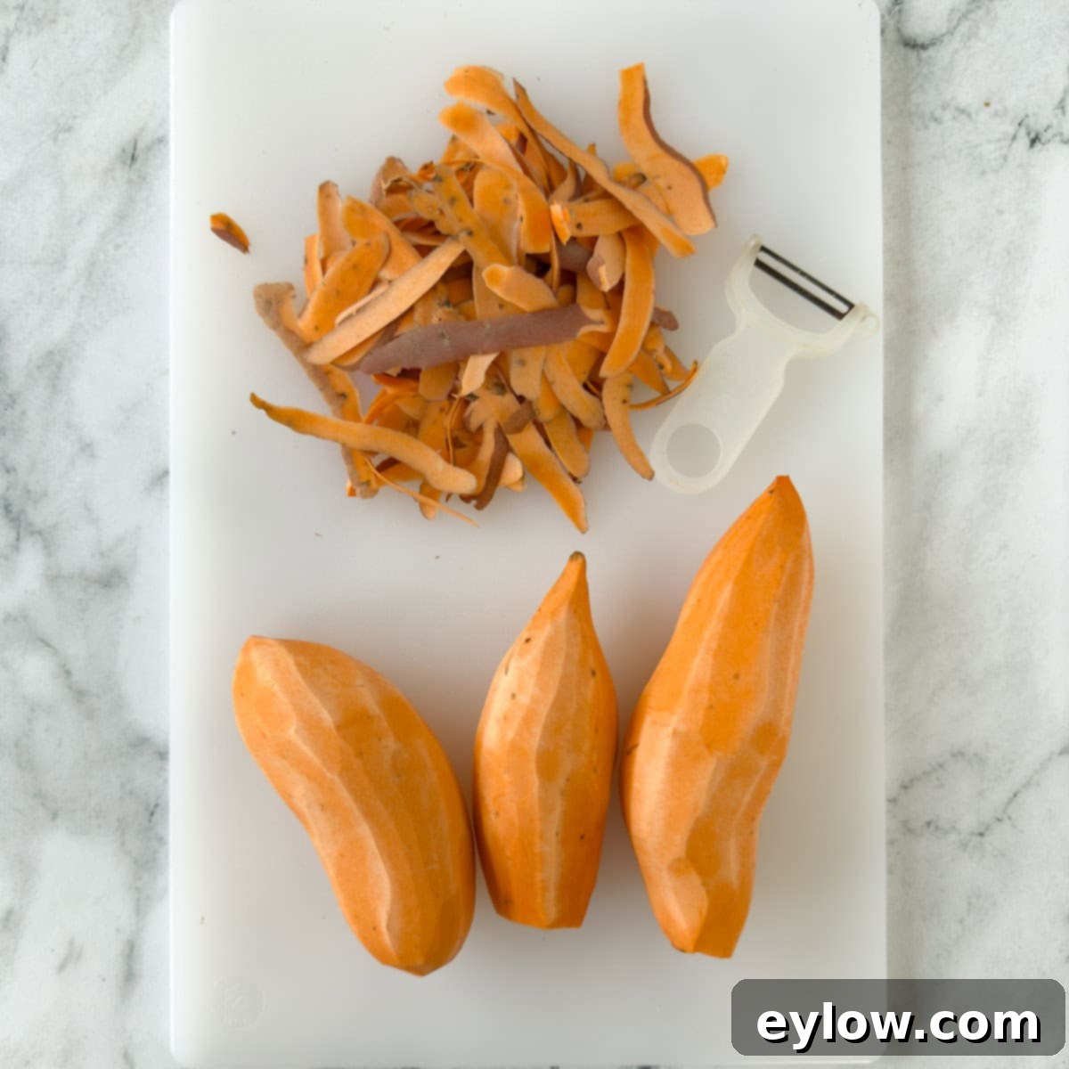 A white cutting board with peeled orange sweet potatoes and their peels. 