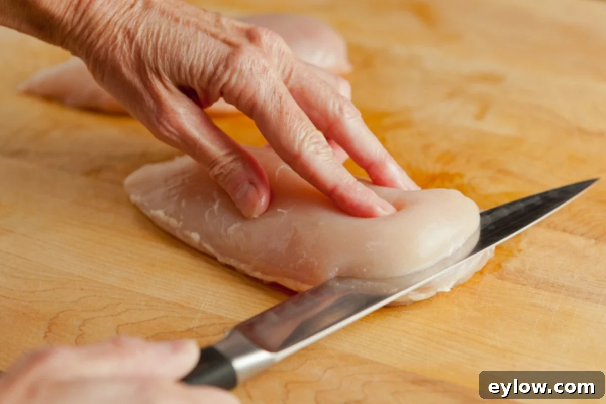 Starting to slice a boneless, skinless chicken breast in half horizontally with a sharp knife on a wood cutting board.