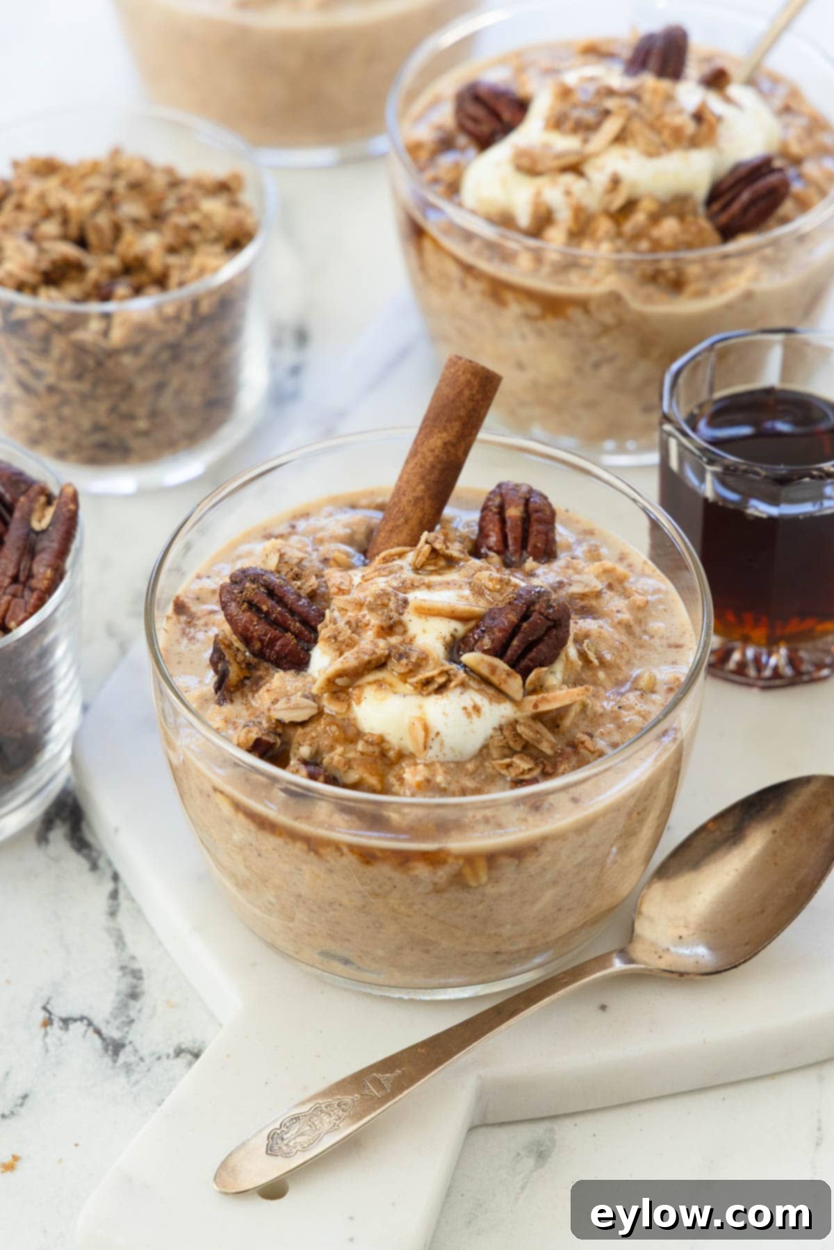Bowls of pumpkin overnight oats with cinnamon stick, pecans, yogurt, and a silver spoon, evoking autumn comfort.