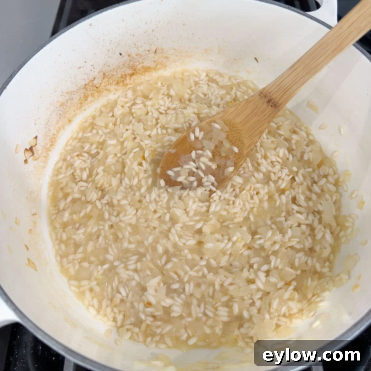 Broth added to simmering rice in a white pan stirring with wooden spoon.