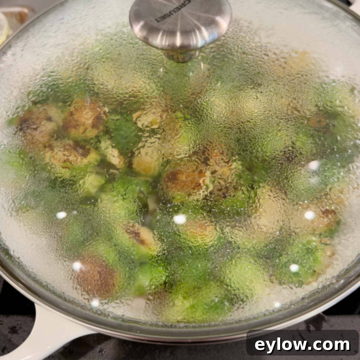 Golden Brussels Sprouts with Crispy Pancetta 10 A wide, shallow pan with a glass lid, actively cooking vibrant green vegetables on a stovetop. Steam can be seen gently escaping, indicating the Brussels sprouts are tenderizing under cover.